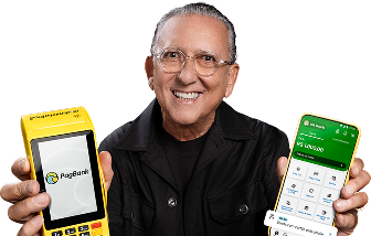 Galvão Bueno segurando uma máquina de cartão do PagBank em uma mão e um smartphone com o aplicativo PagBank aberto na outra, sobre fundo amarelo.