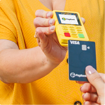 Uma pessoa segura uma maquininha do PagBank enquanto outra aproxima um cartão Visa para realizar um pagamento.